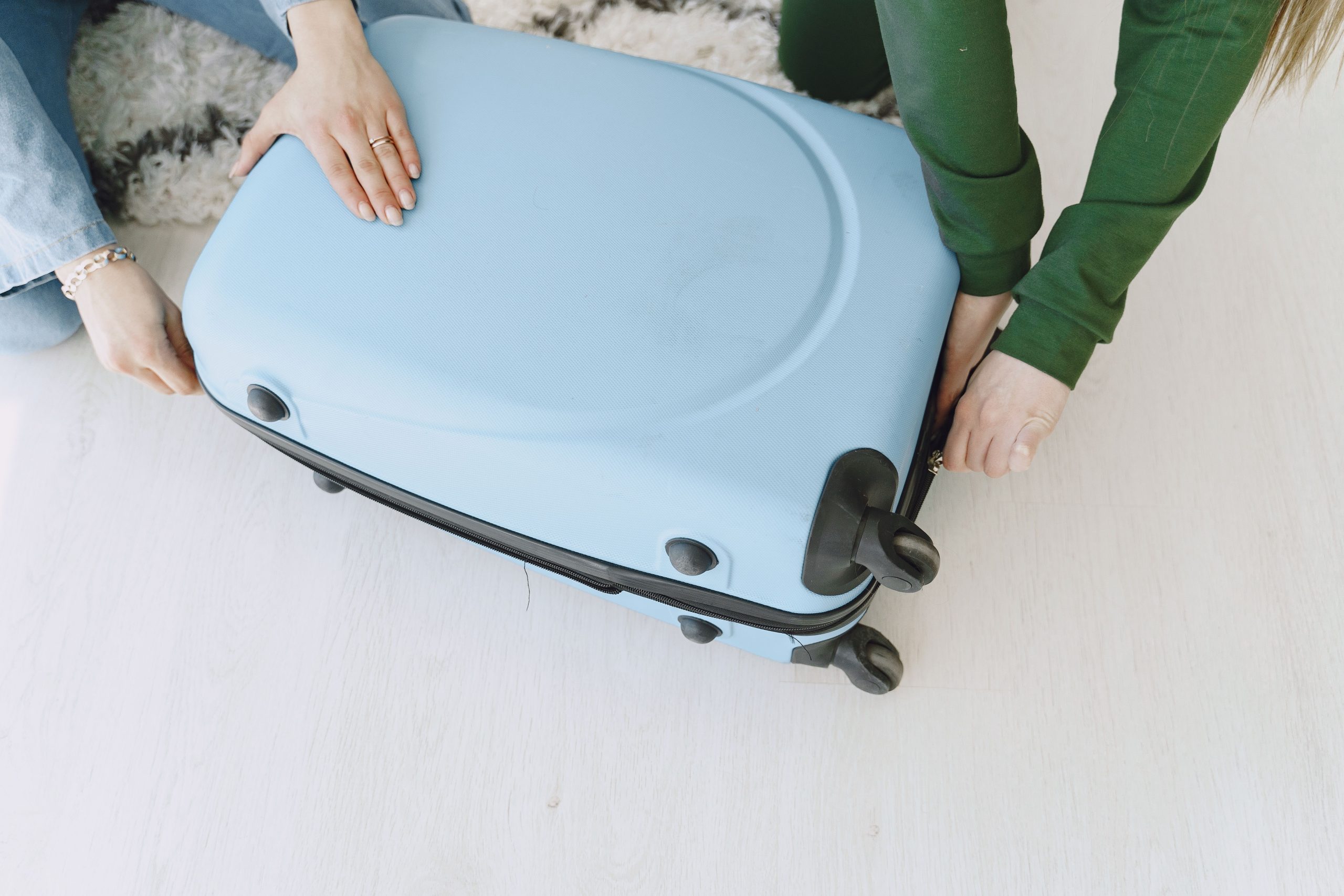 Unrecognizable female friends in casual clothes sitting on floor with fluffy carpet and opening suitcase together while preparing for journey and adventure